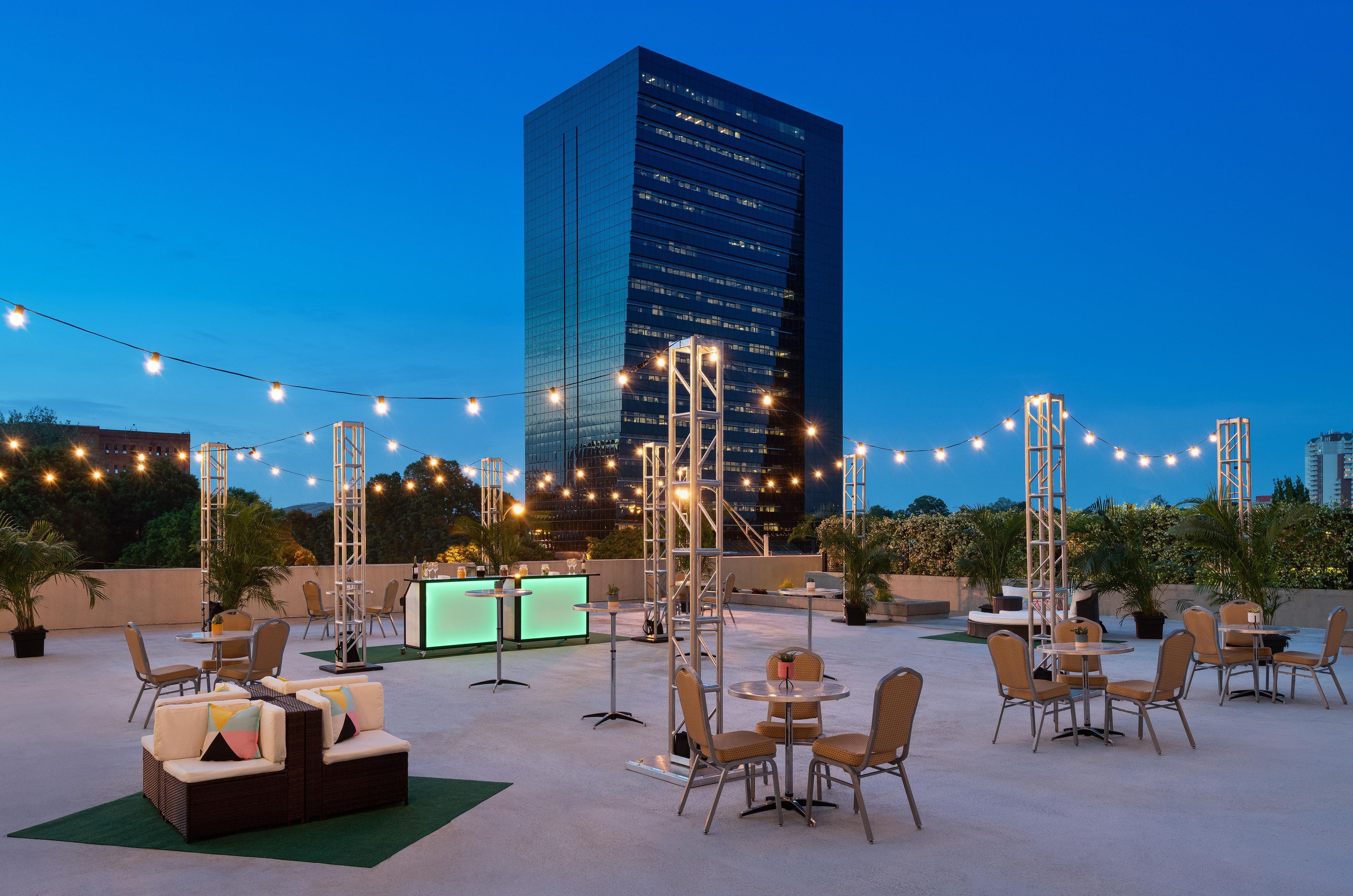 An outdoor seating area with tables and chairs, a bar, and string lights against a city skyline at dusk.