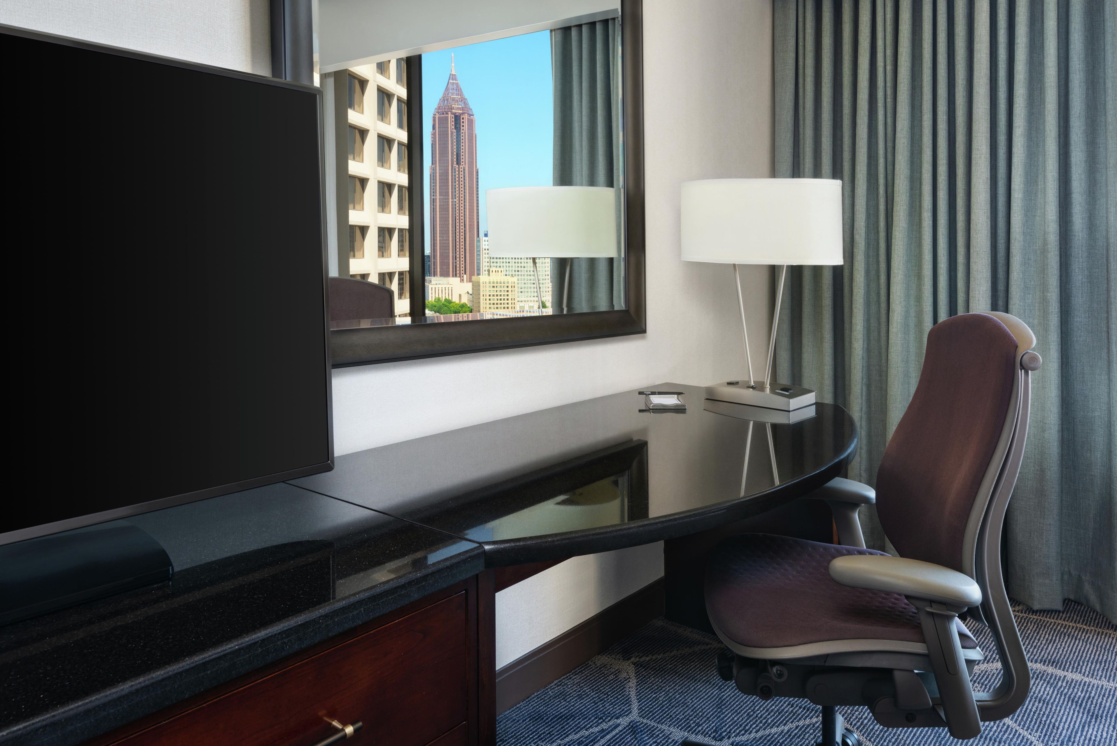 A hotel room with a large flat-screen TV, a desk with a chair, a lamp, and a view of a cityscape.
