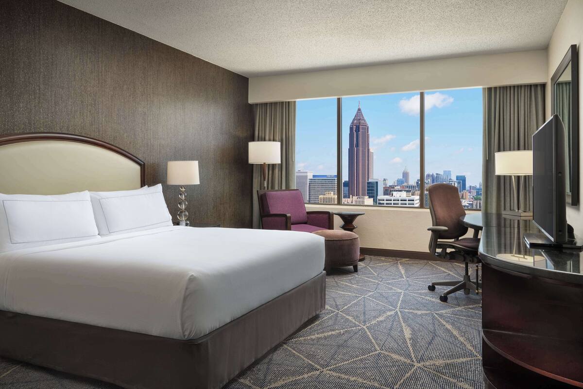 A hotel room with a large bed, two chairs, a desk, and a TV. The room has a city view with a tall building in the background.