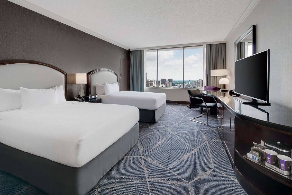 A hotel room with two beds, a desk, a TV, and a large window with a city view.