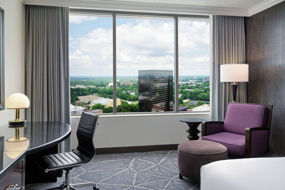 A hotel room with a large window, a round table, a purple armchair, a small ottoman, and a lamp.