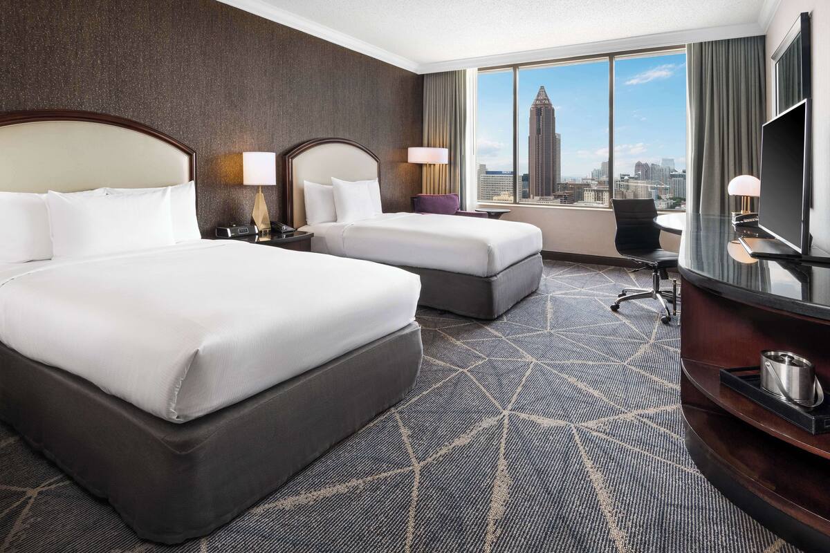A hotel room with two beds, a desk, a chair, a TV, and a view of a cityscape.