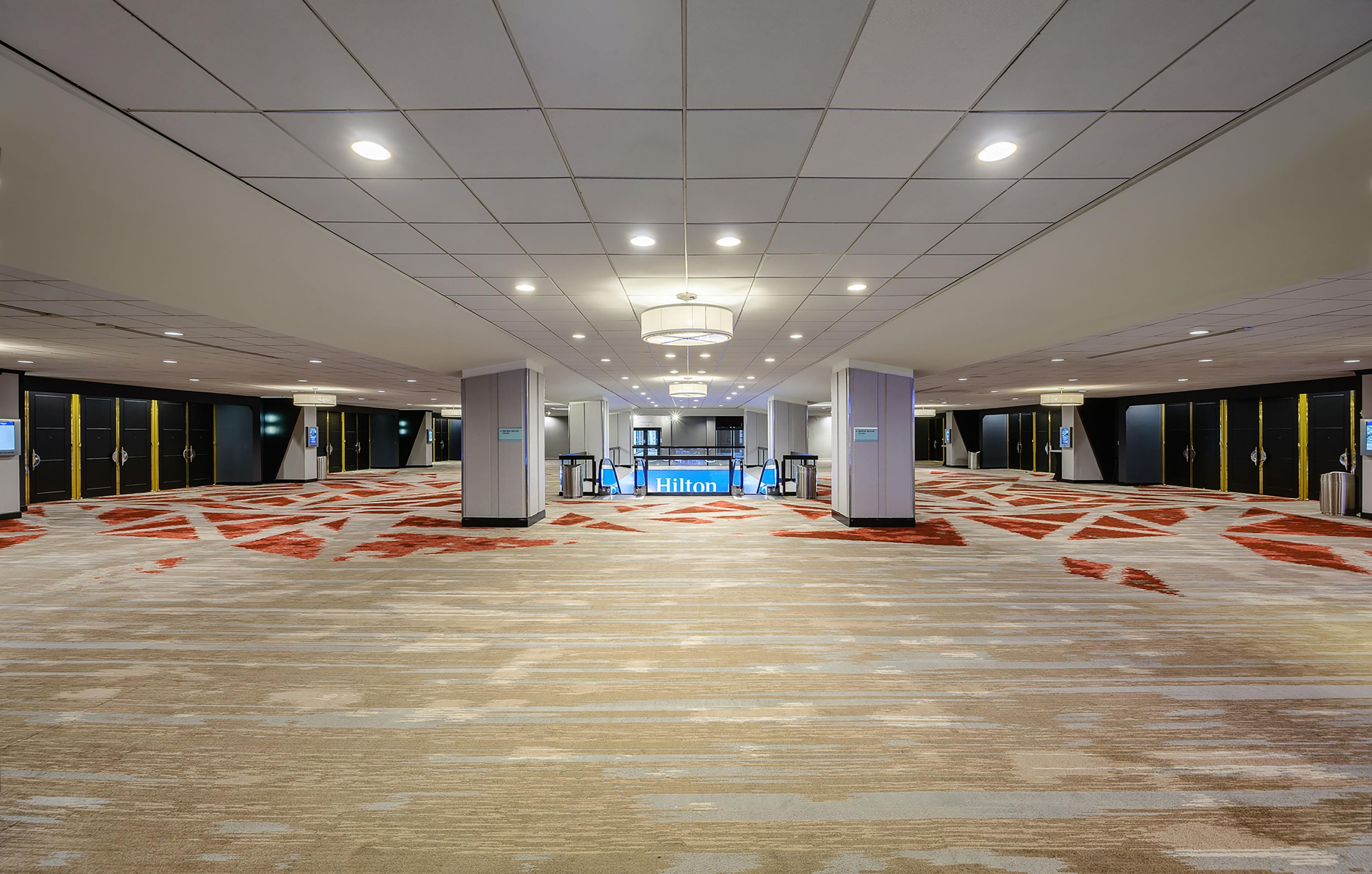 A spacious hotel lobby with a high ceiling, recessed lighting, and a central reception desk.
