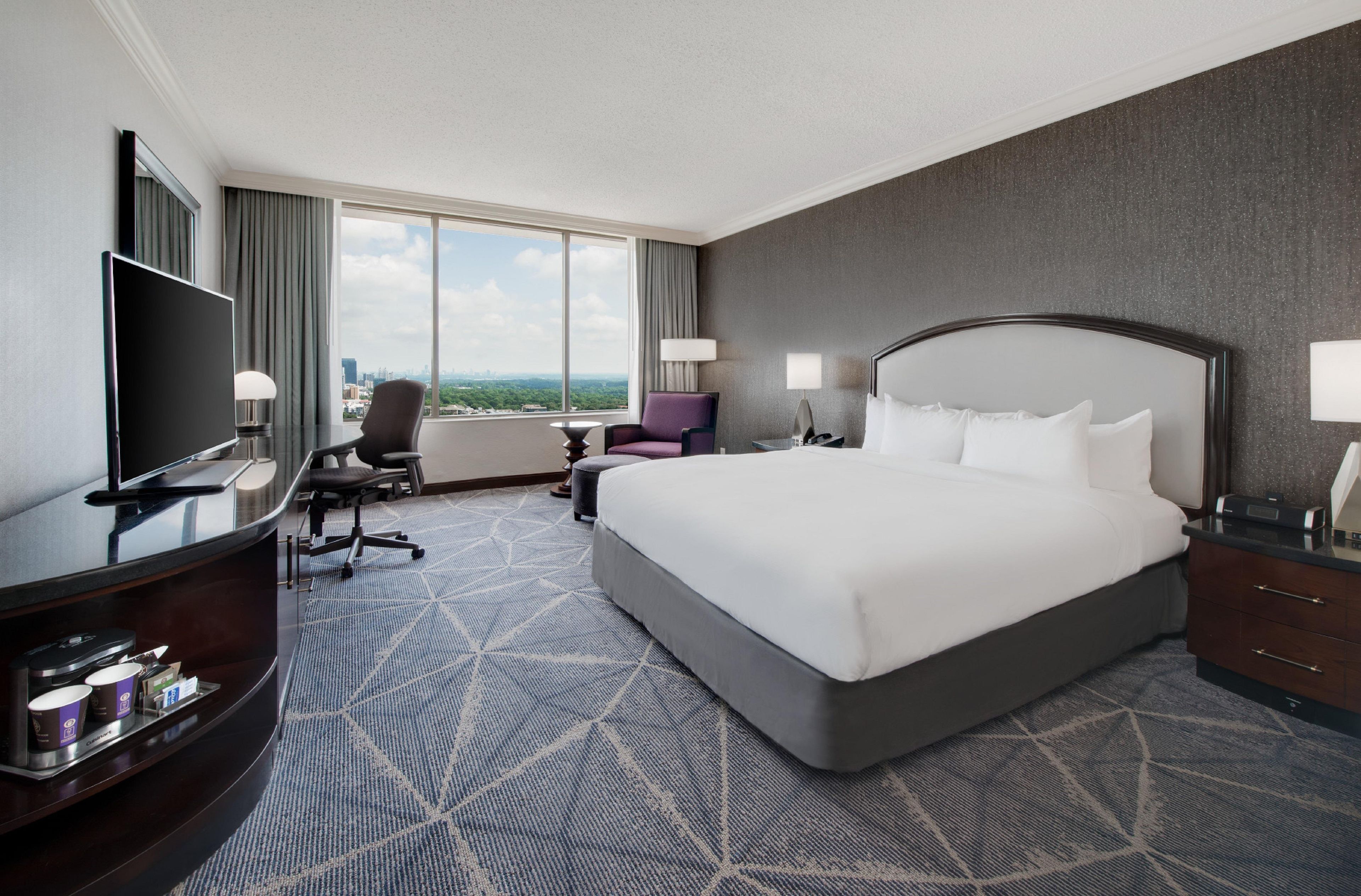 A hotel room with a large bed, a desk with a computer, a TV, and a view of the city.