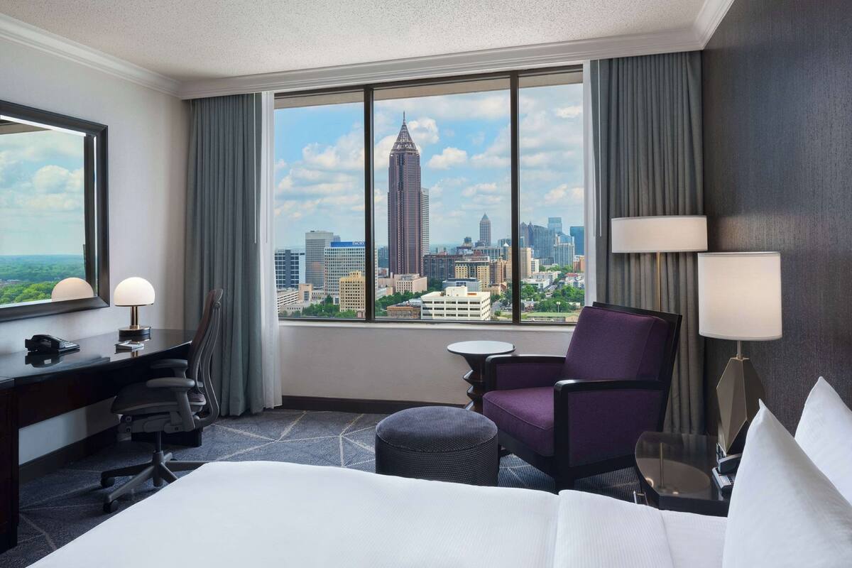 A hotel room with a large bed, a desk with a lamp, a chair, a small table, and a view of a city skyline.