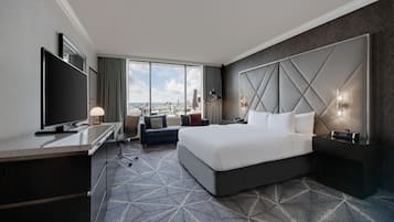 A hotel room with a large bed, a desk with a flat-screen TV, a sofa, and a city view.
