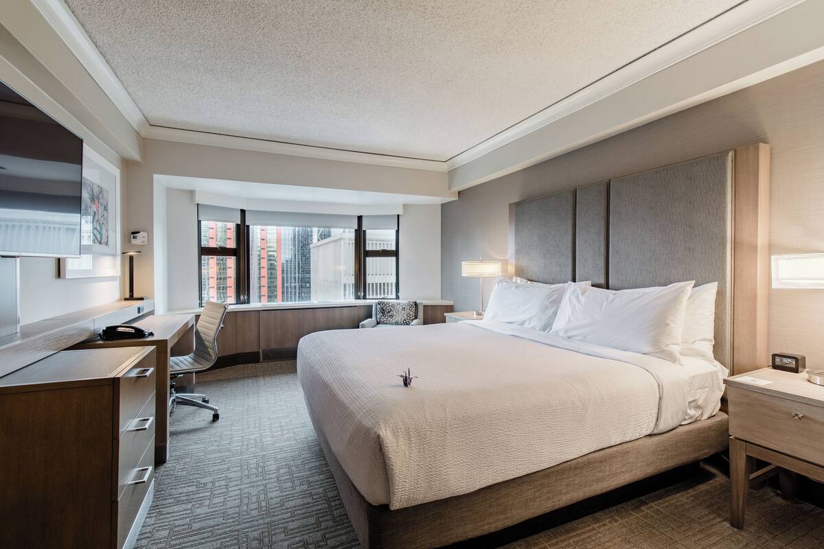 A hotel room with a large bed, a desk, a chair, a nightstand, and a window with a view of buildings.