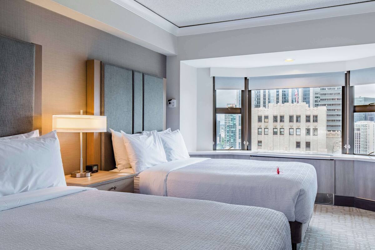 A hotel room with two beds, a nightstand, a lamp, and a large window offering a city view.