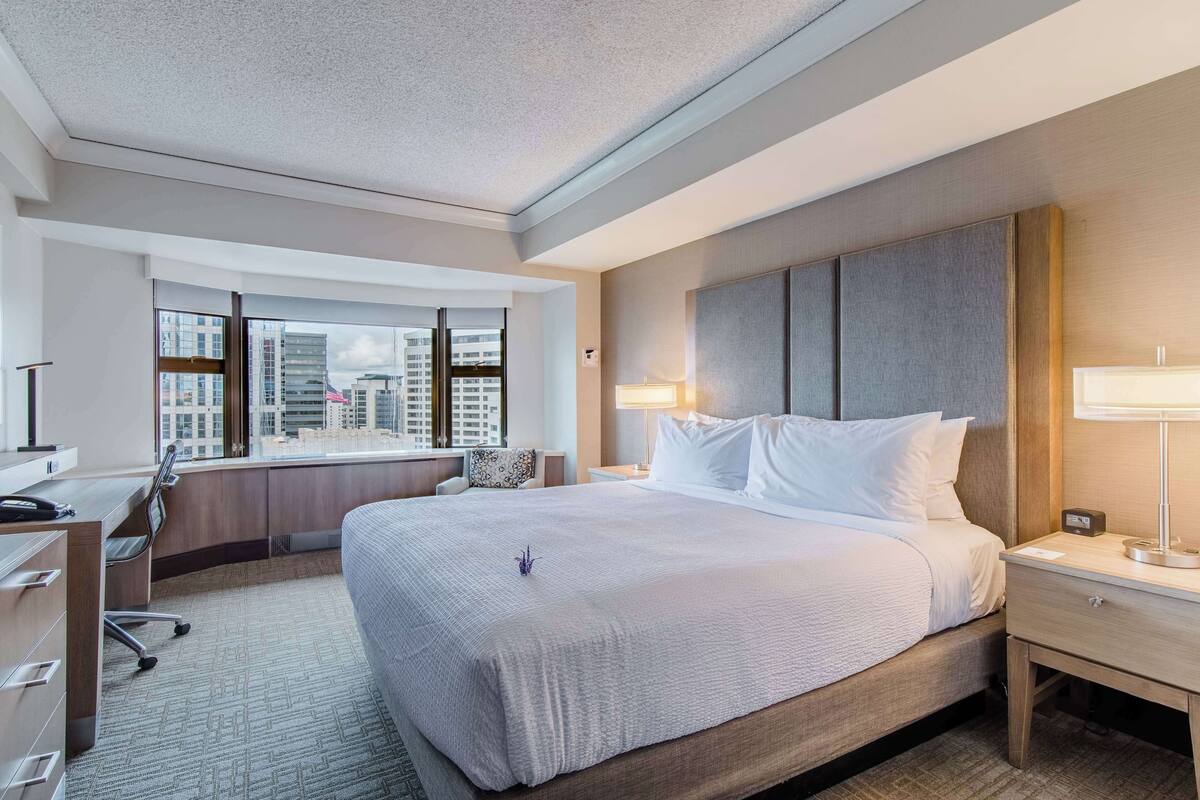 A modern hotel room with a large bed, a desk, and a view of the city through large windows.