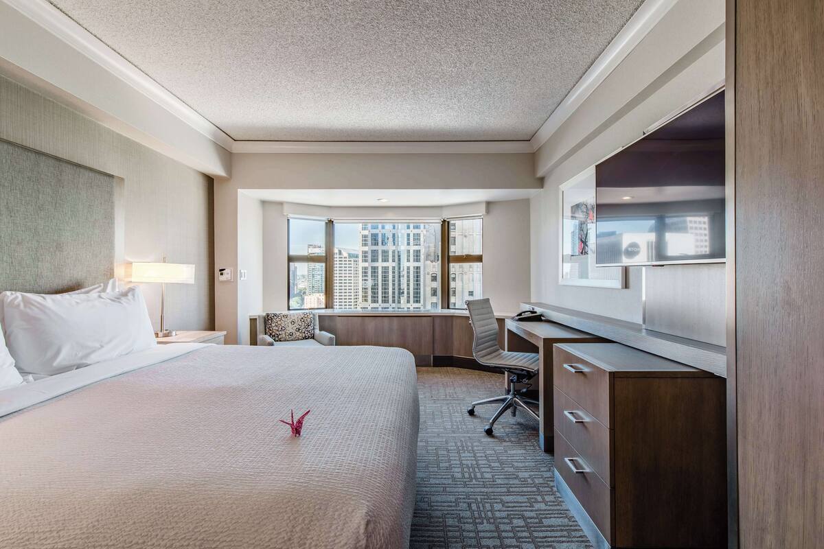 A hotel room with a large bed, a desk, a chair, a TV, and a view of the city through large windows.
