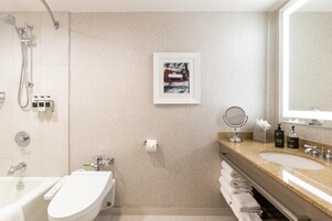 A modern bathroom with a toilet, bidet, and shower. There is a framed picture on the wall and a sink with toiletries.