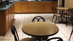 Ein kleines, helles Café mit Holzausbau, einer Theke mit verschiedenen Speisen, einer Kaffeemaschine und einer Wanduhr.