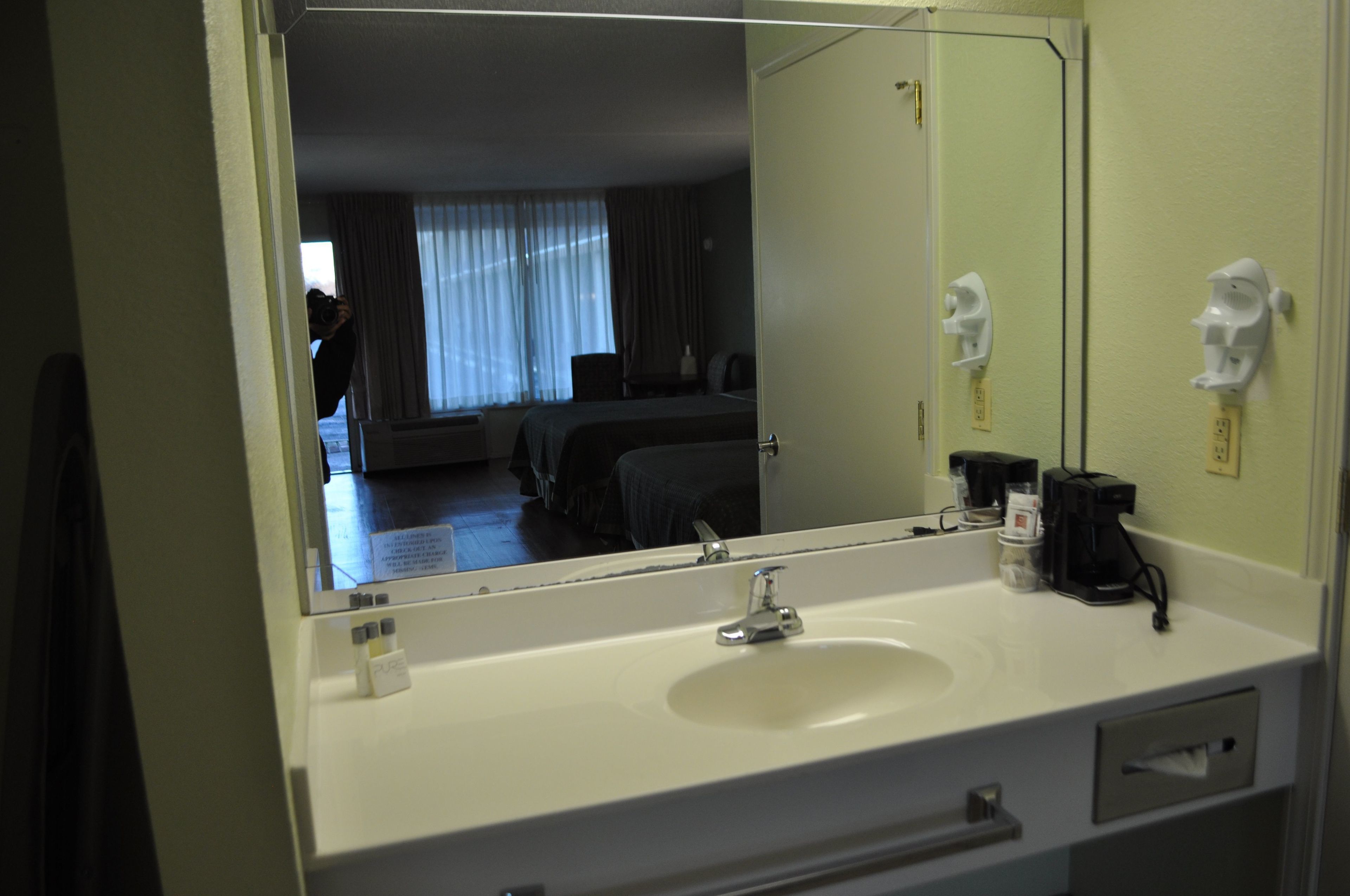 A hotel bathroom with a large mirror, a sink, a coffee maker, and a towel dispenser.