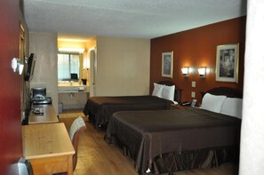 A hotel room with two beds, a desk, a chair, a small kitchenette, and a bathroom visible through an open door.