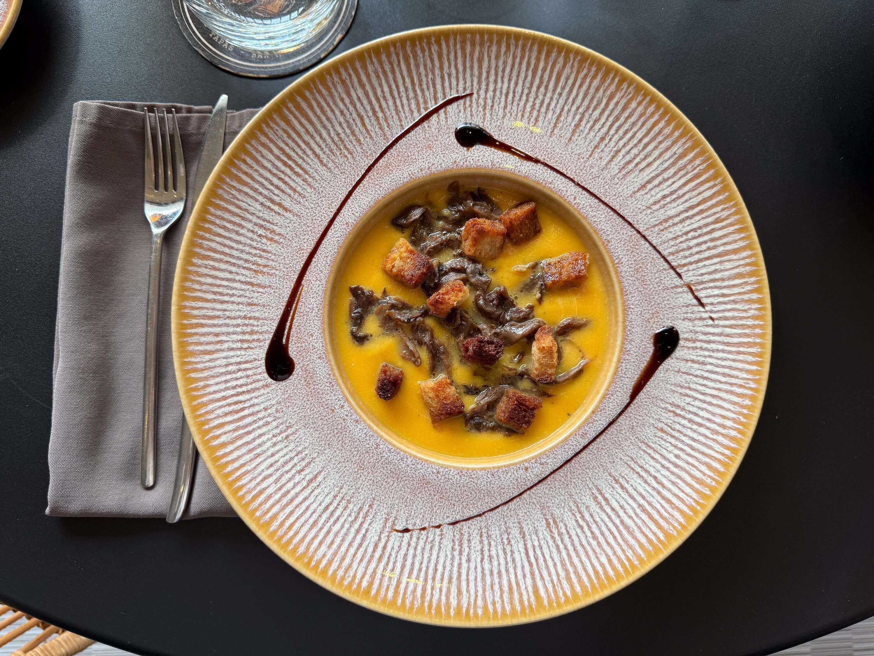 A bowl of soup with croutons and a drizzle of sauce on a textured plate.