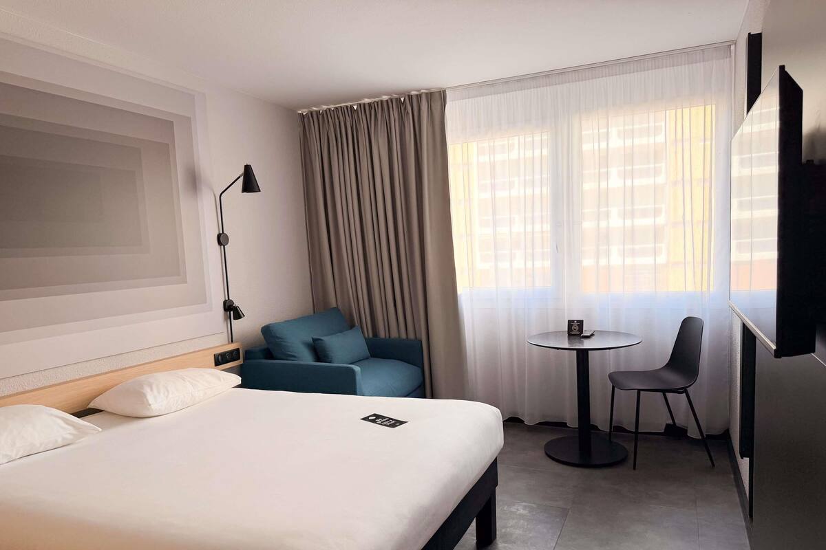 A hotel room with a bed, a blue armchair, a small round table with a chair, a TV, and a window with curtains.