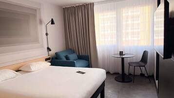 Quarto de hotel com uma cama, uma poltrona azul, uma mesa redonda pequena com uma cadeira, uma televisão e uma janela com cortinas.