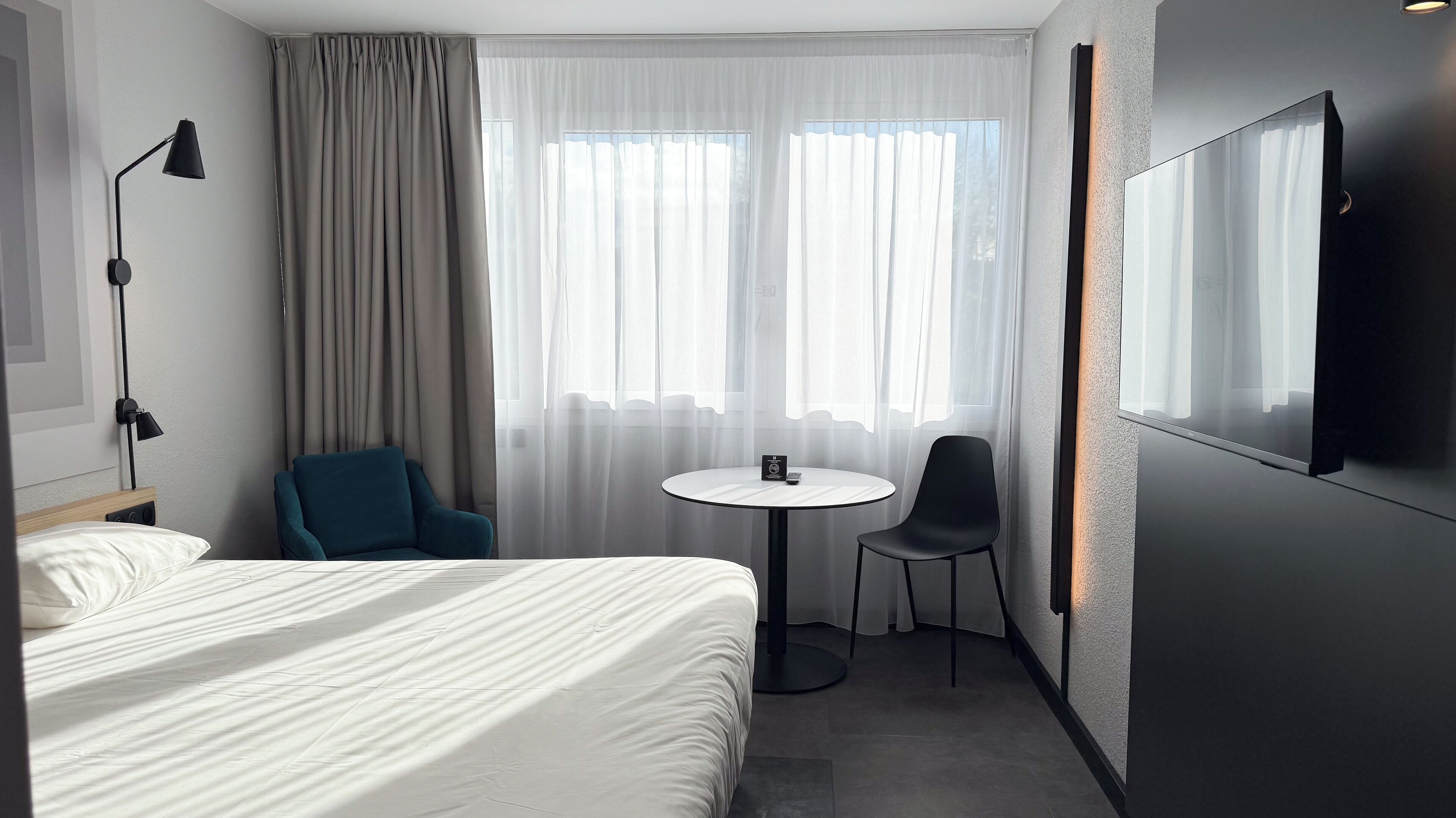Ett hotellrum med en säng, ett skrivbord, en stol, ett runt bord, en TV och tunna, genomskinliga gardiner.