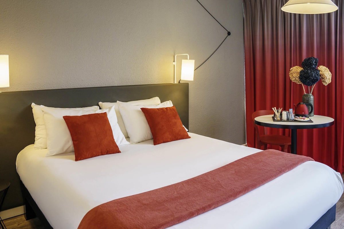 A hotel room with a large bed, a bedside table with a lamp, and a view of the outside through red curtains.