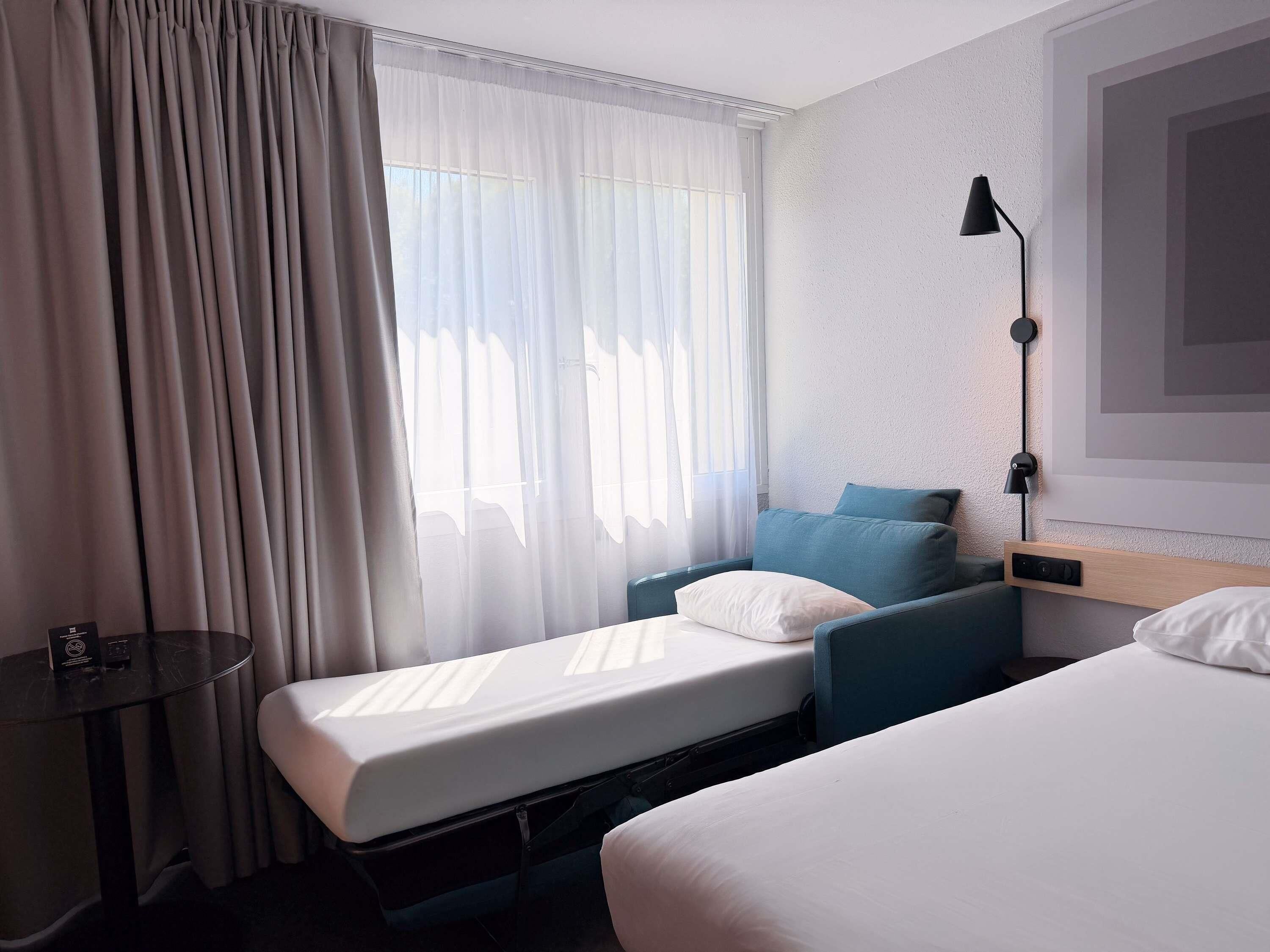 A hotel room with two beds, a bedside table, a lamp, and a window with curtains.