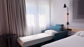 A hotel room with two beds, a bedside table, a lamp, and a window with curtains.