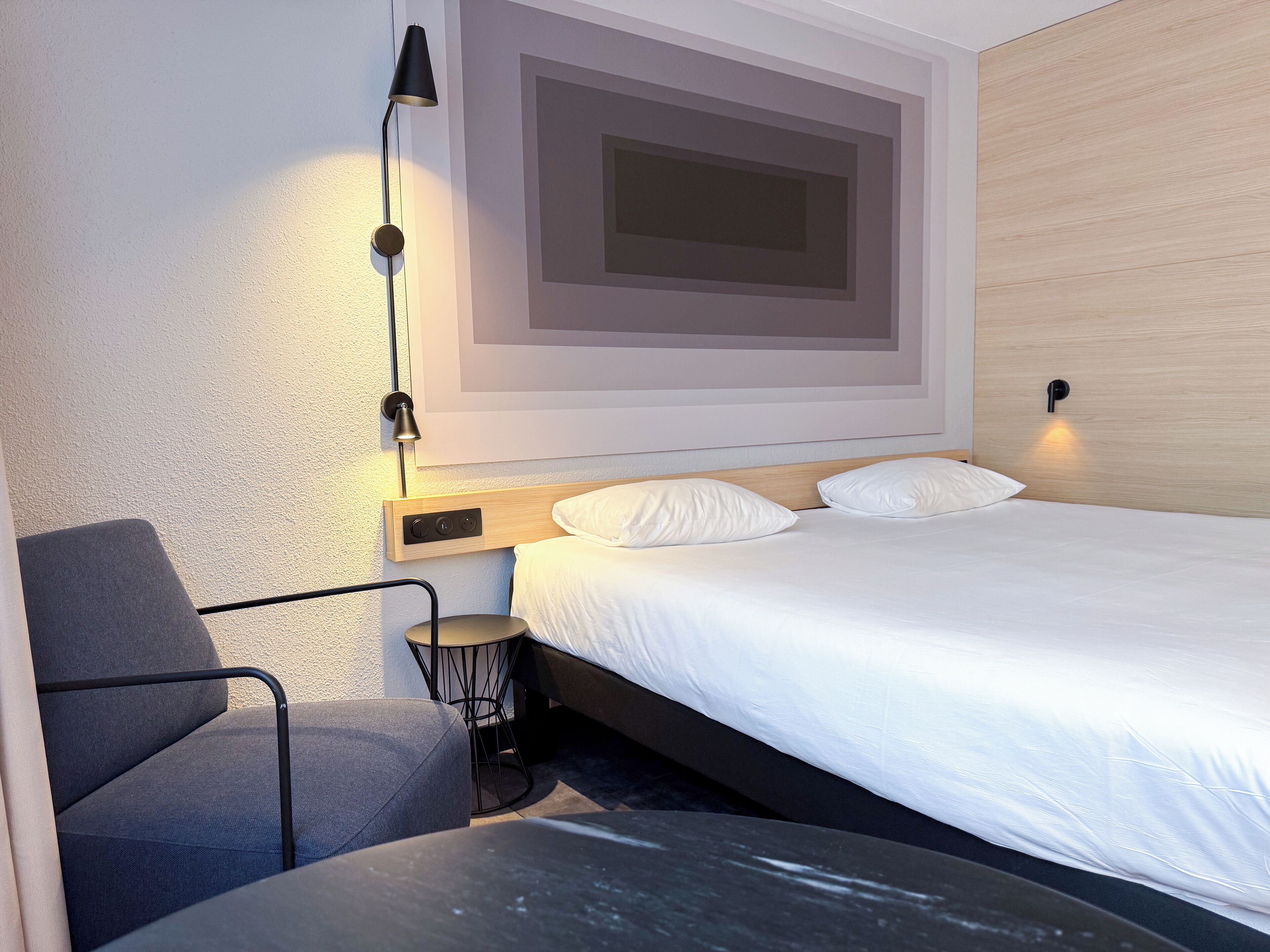 A modern hotel room with a bed, a desk, a chair, and a wall-mounted lamp.