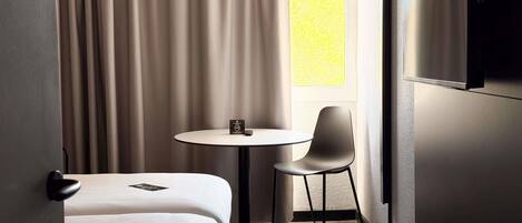 A hotel room with a bed, a round table with a chair, a TV mounted on the wall, and a window with curtains.