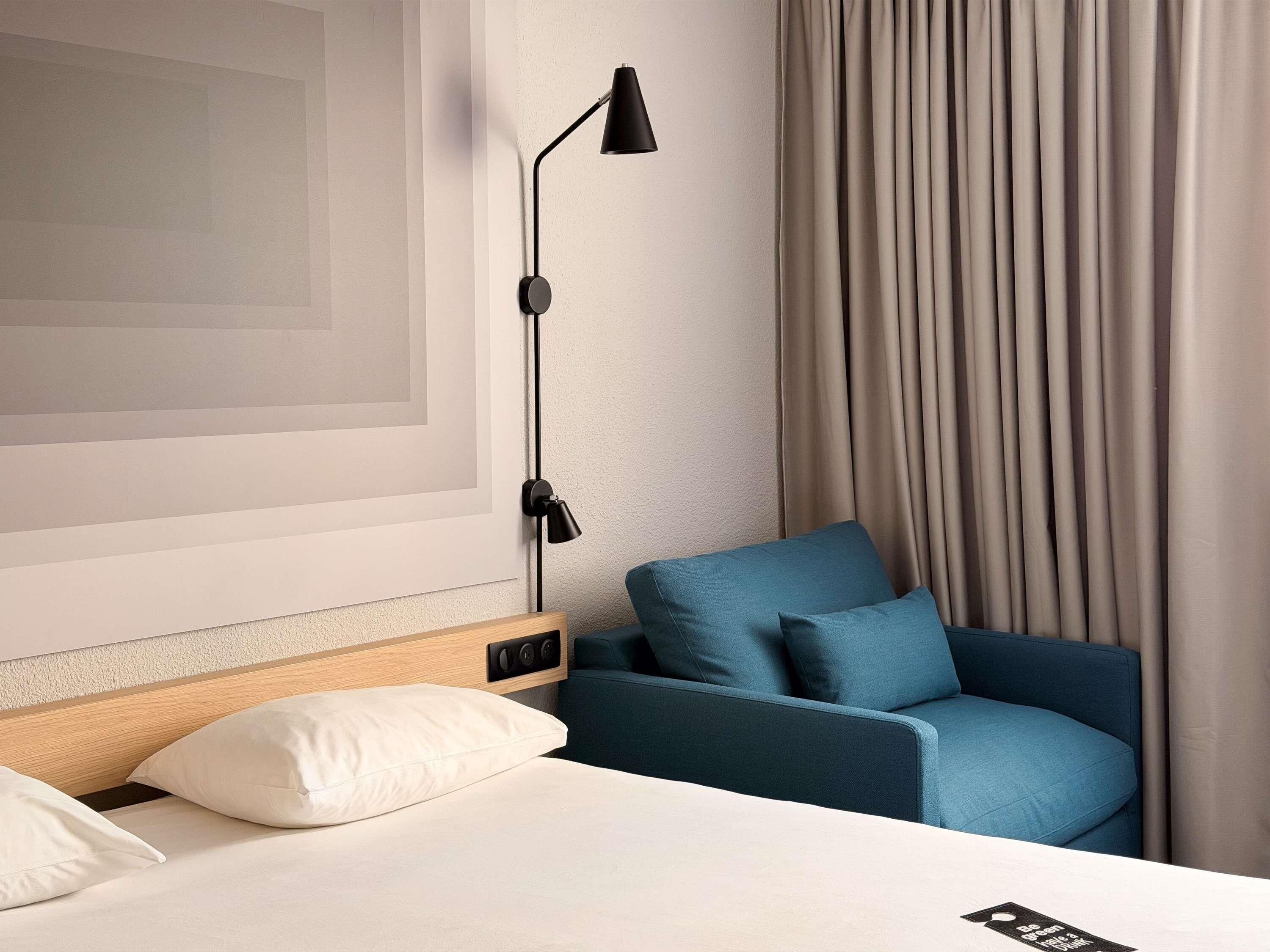 A hotel room with a bed, a blue armchair, a wall-mounted lamp, and a large window with curtains.