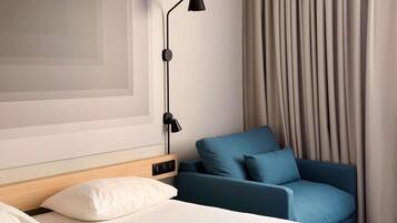 A hotel room with a bed, a blue armchair, a wall-mounted lamp, and a large window with curtains.