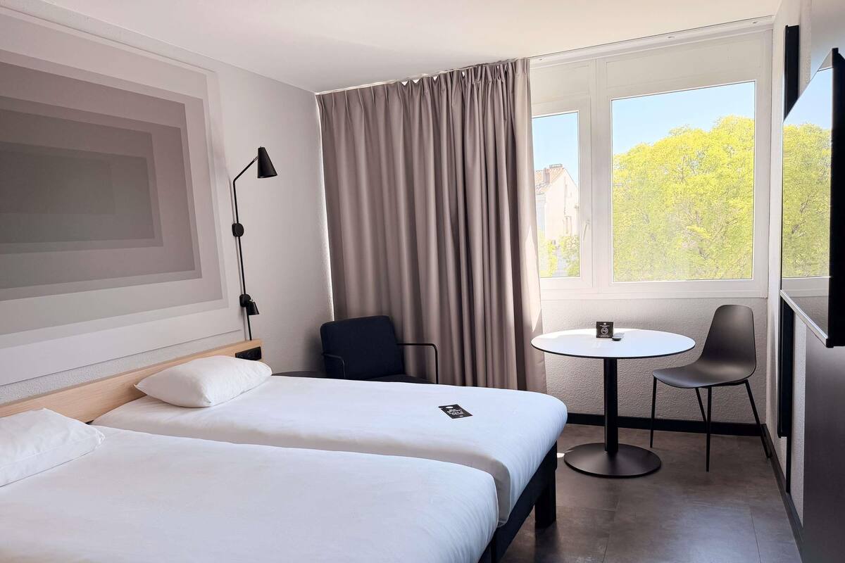 A hotel room with two beds, a desk, a chair, a lamp, and a window with a view of trees.