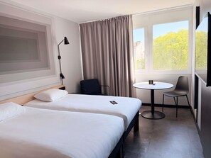 A hotel room with two beds, a desk, a chair, a lamp, and a window with a view of trees.
