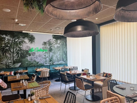 A restaurant with a mural of tropical foliage and a neon sign reading 'Cebena.' Tables are set with glasses and cutlery, and there are hanging pendant lights.. ibis Ales Centre Ville Hotel