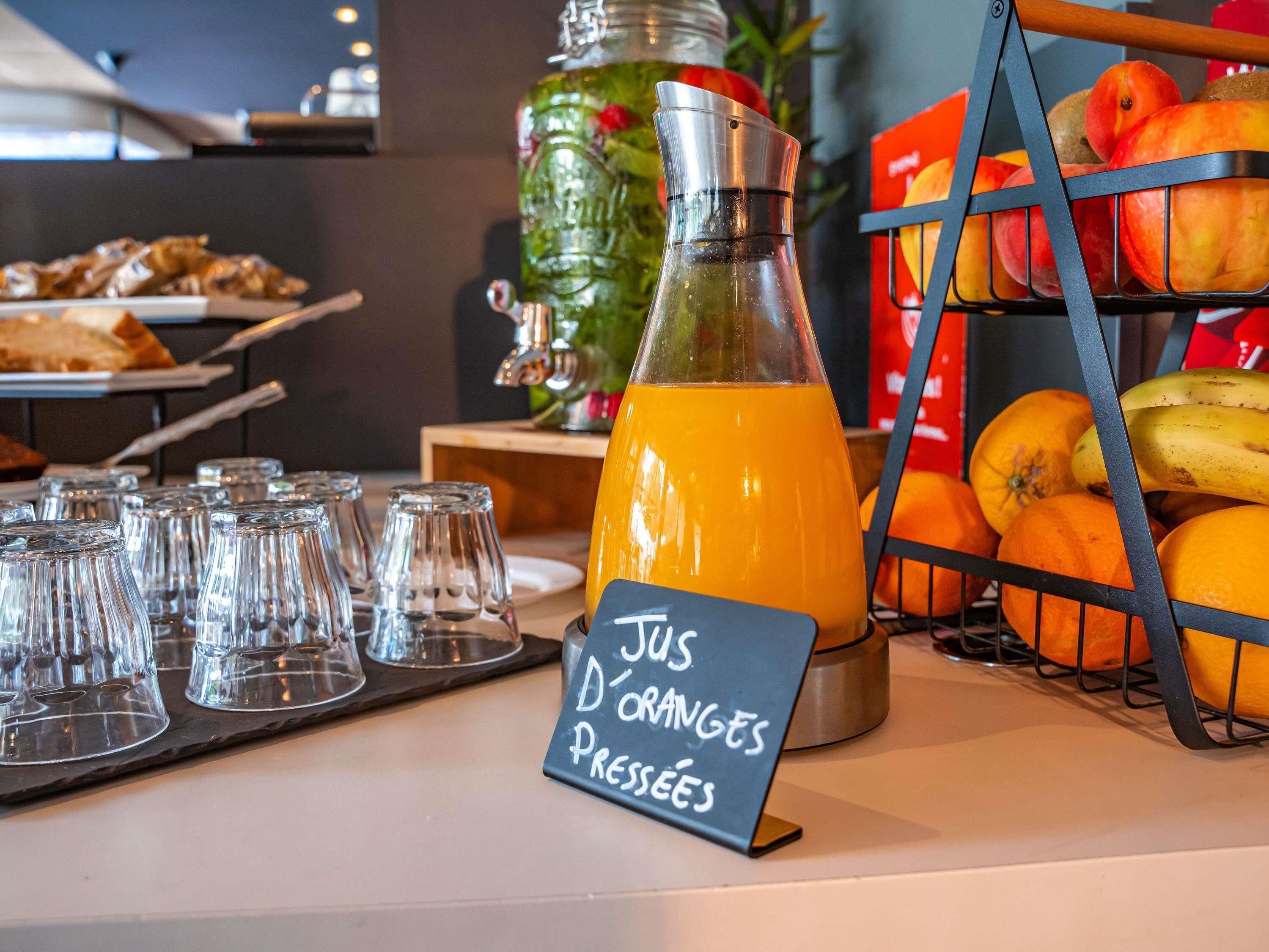 A juice dispenser with a sign indicating 'Jus D'Orange Pressées' and a fruit display with oranges and bananas.