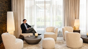 A modern lounge area with a man using a laptop, surrounded by plush armchairs and round tables, with large windows and curtains. - Best Western Plus Delta Park Hotel (Mannheim)