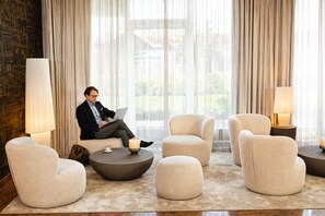 A modern lounge area with a man using a laptop, surrounded by plush armchairs and round tables, with large windows and curtains. - Best Western Plus Delta Park Hotel (Mannheim)