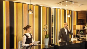 A hotel reception desk with a modern design, a receptionist, and a man on the phone. - Best Western Plus Delta Park Hotel (Mannheim)