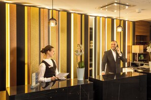 A hotel reception desk with a modern design, a receptionist, and a man on the phone. - Best Western Plus Delta Park Hotel (Mannheim)