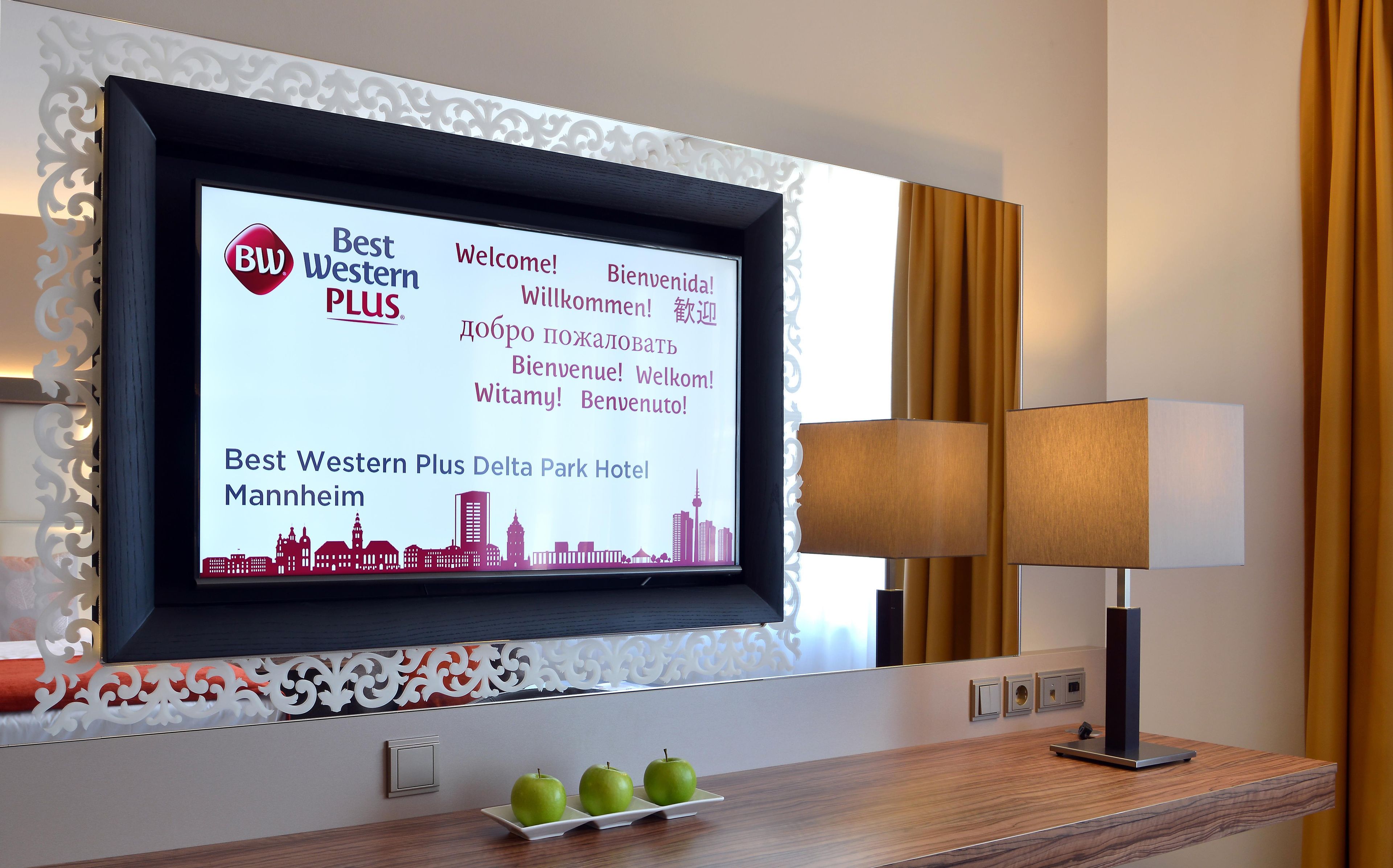 Ein Hotel-Rezeptionsbereich mit einem digitalen Willkommensbildschirm, der den Namen und den Standort des Hotels anzeigt, einer dekorativen Wandverkleidung, einem Tisch mit Äpfeln und einer Lampe.