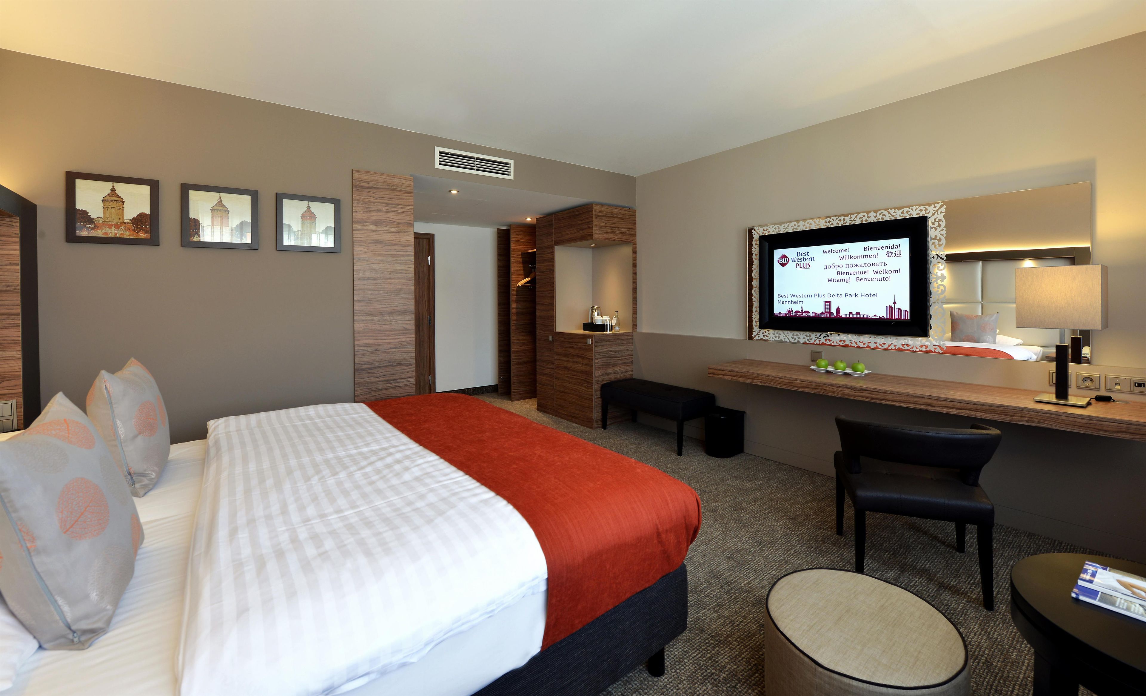 Een hotelkamer met een bed, een bureau, een stoel, een klein krukje, een televisie en een spiegel.