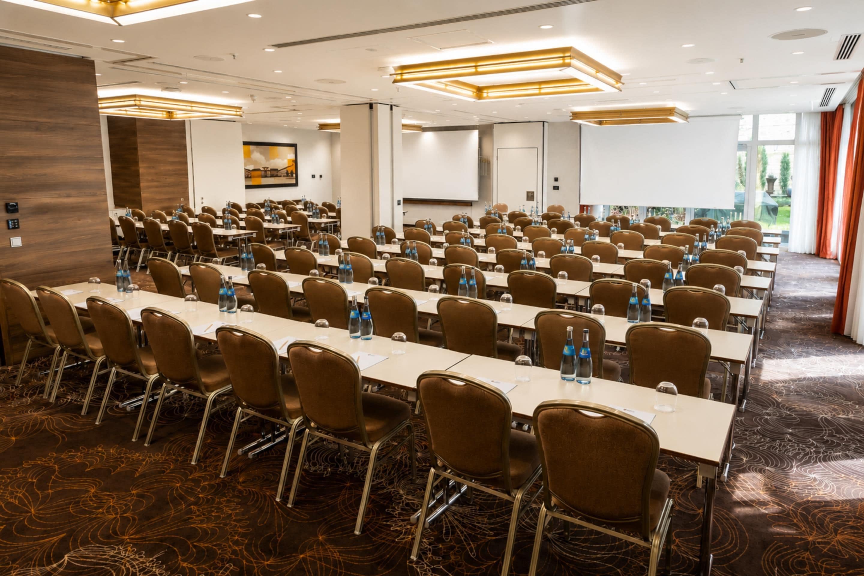 A conference room with rows of tables and chairs, equipped with water bottles, and a large screen at the front.
