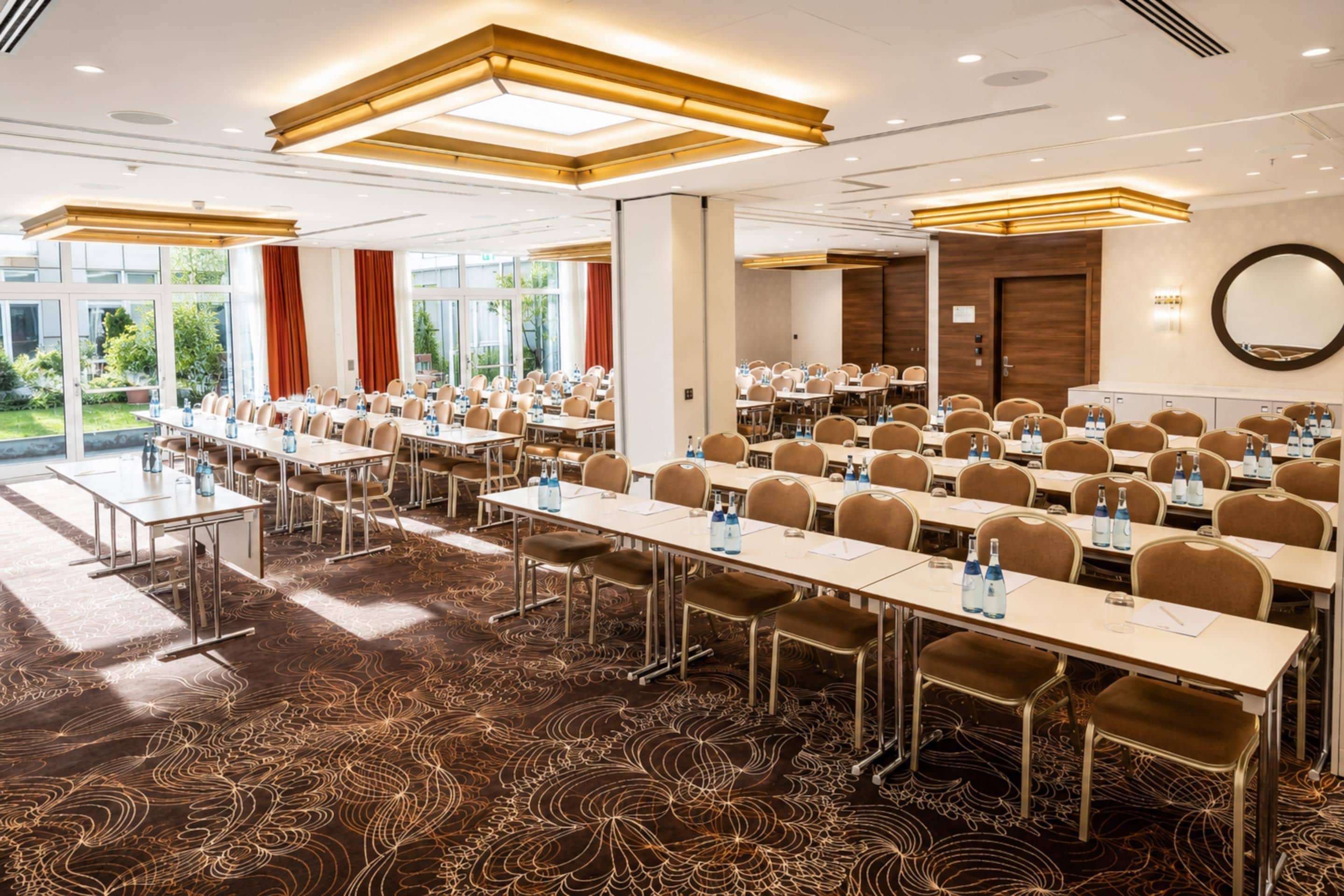 A conference room with long tables and chairs, equipped with water bottles, in a hotel with a patterned carpet and large windows.