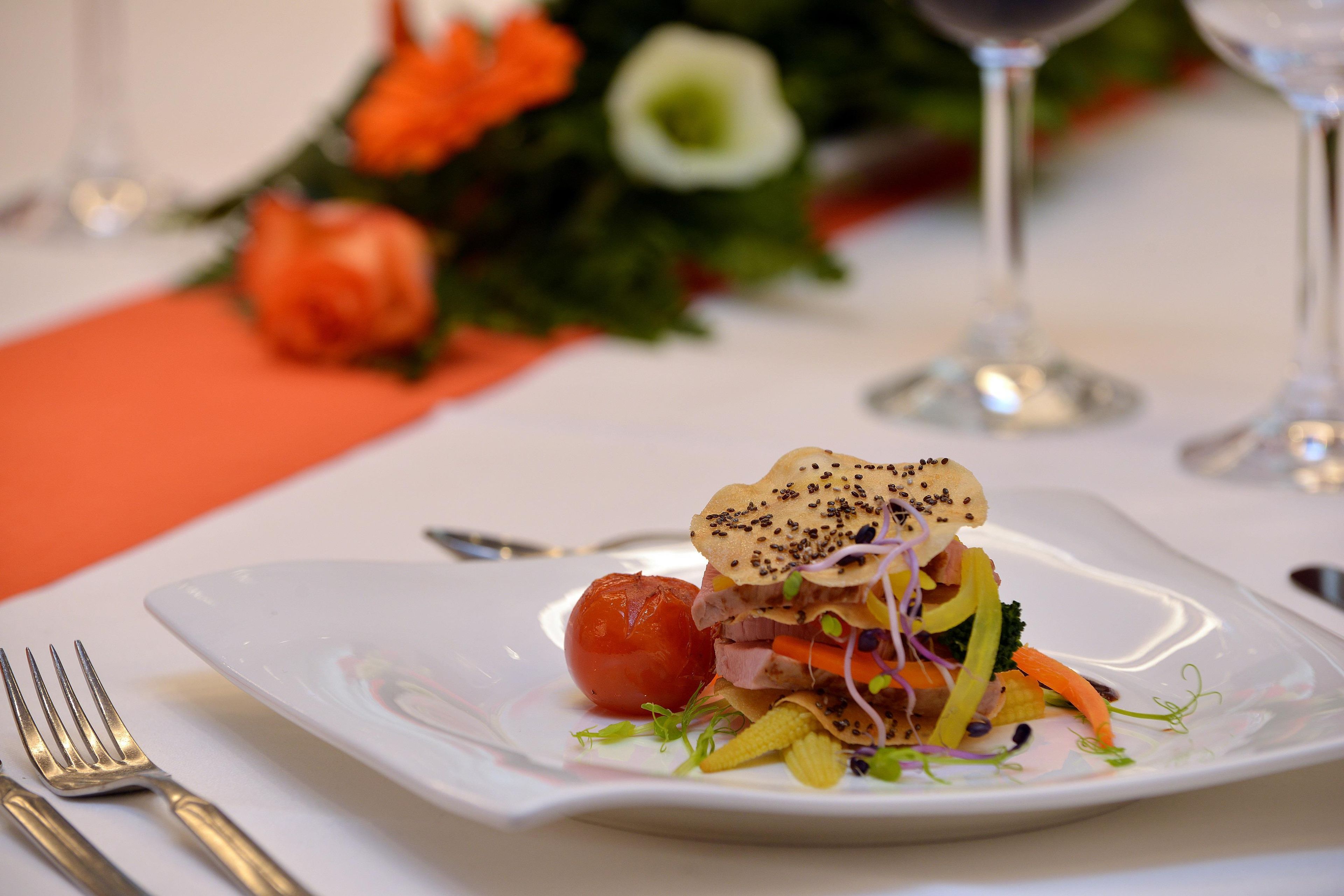 Un plato cuadrado con ensalada, un tomate pequeño y un adorno sobre un mantel blanco.
