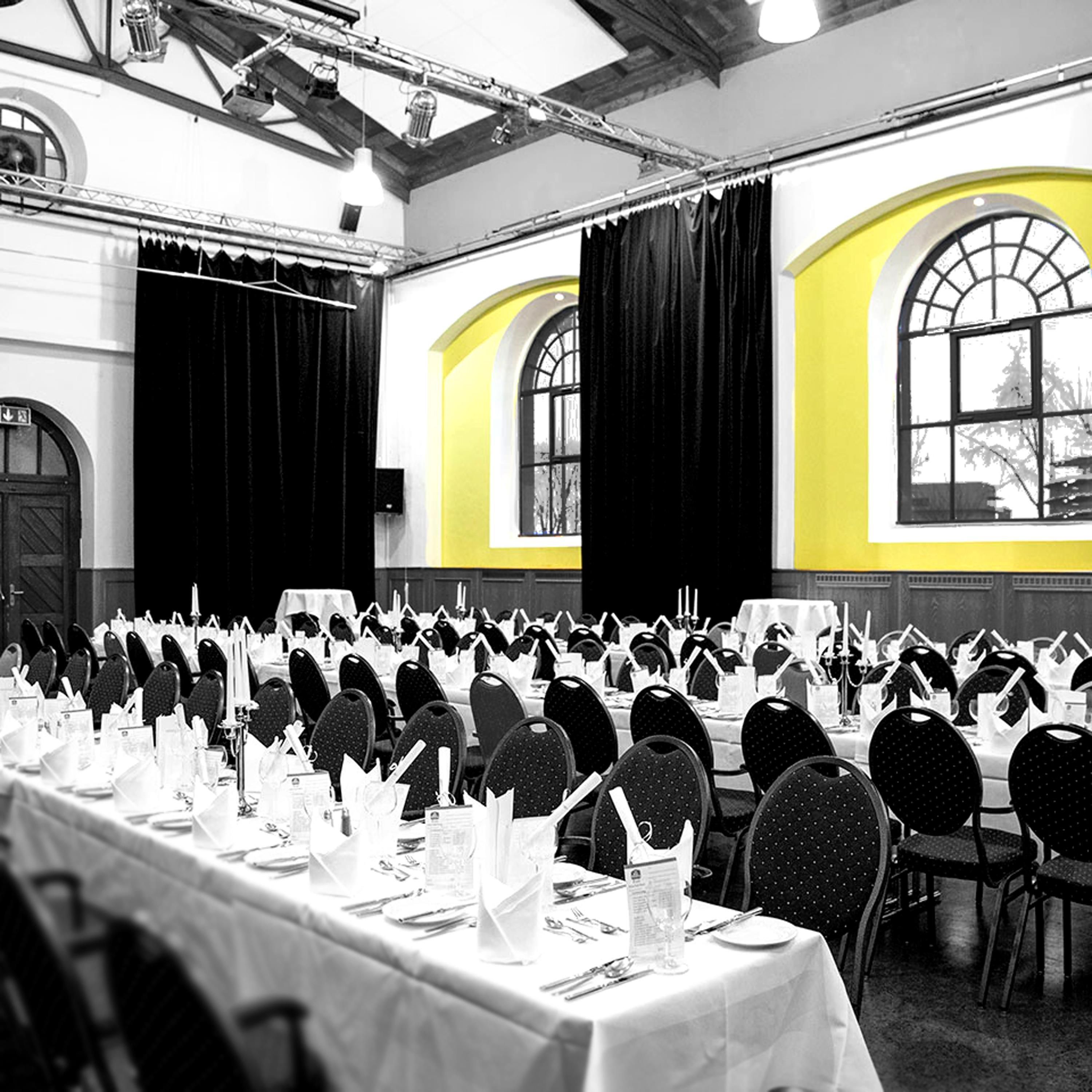 A banquet hall with rows of tables set for a formal event, large windows with arched tops, and a high ceiling with exposed beams.