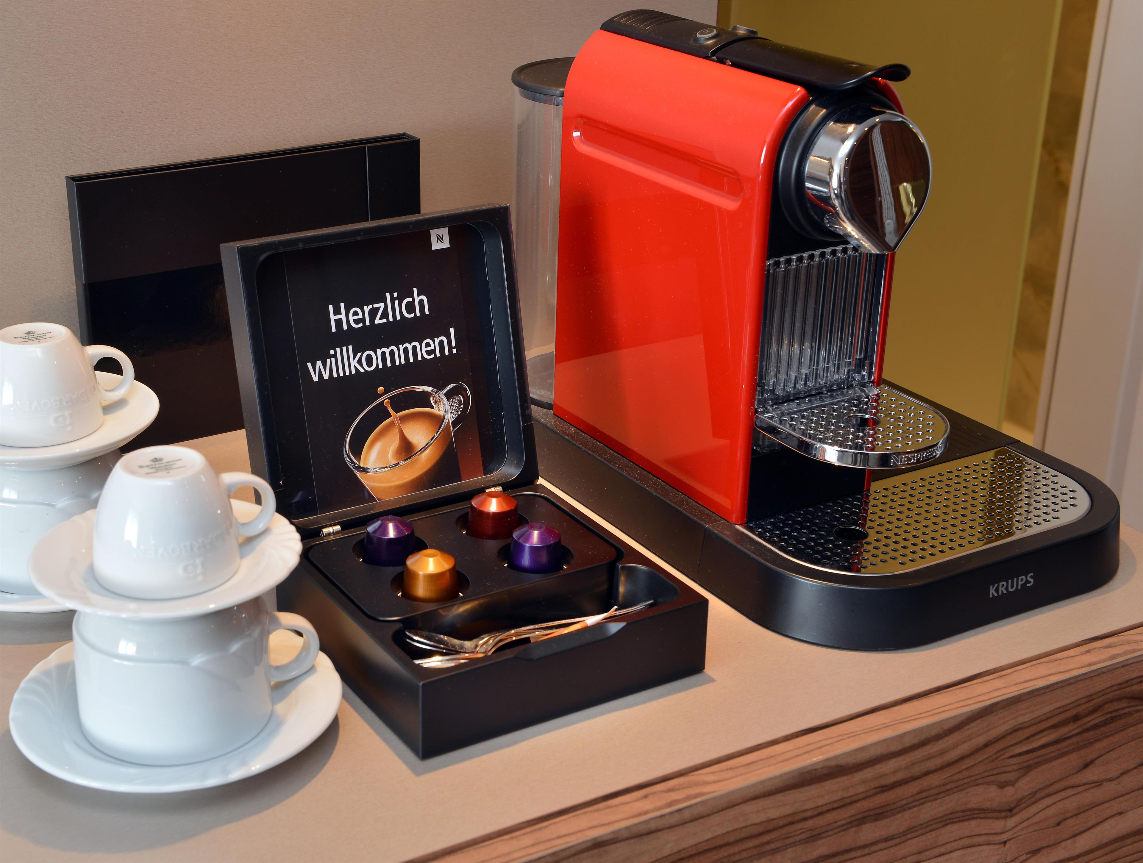Une machine à café avec une base rouge et un bac d’égouttement noir, accompagnée d’une boîte contenant quatre capsules et une cuillère.
