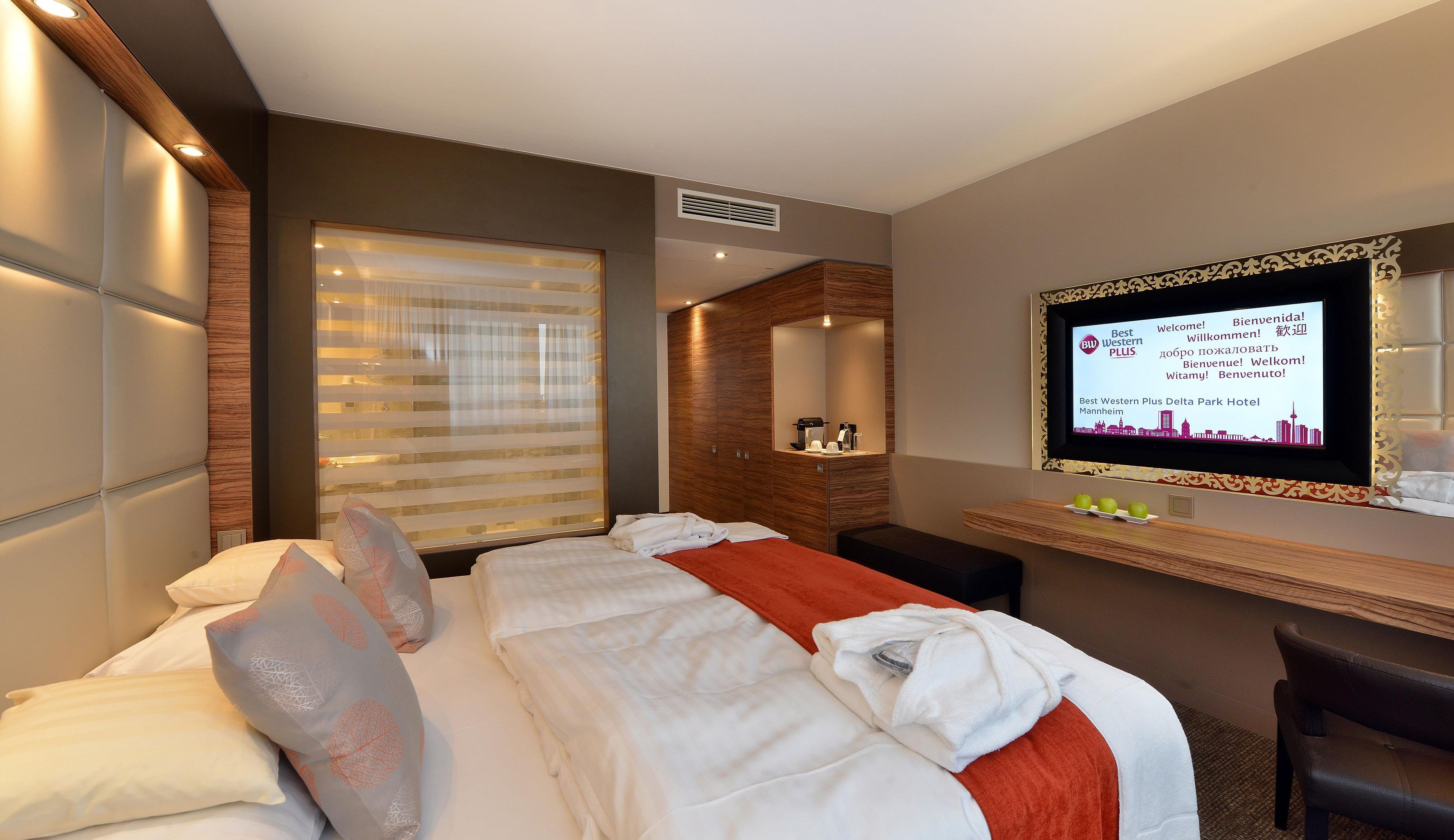 A hotel room with a bed, a TV, a desk, and a window with blinds.