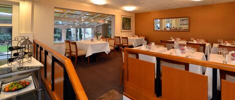 A dining area with multiple tables set for service, a staircase with a railing, and a view of the outdoors through large windows.