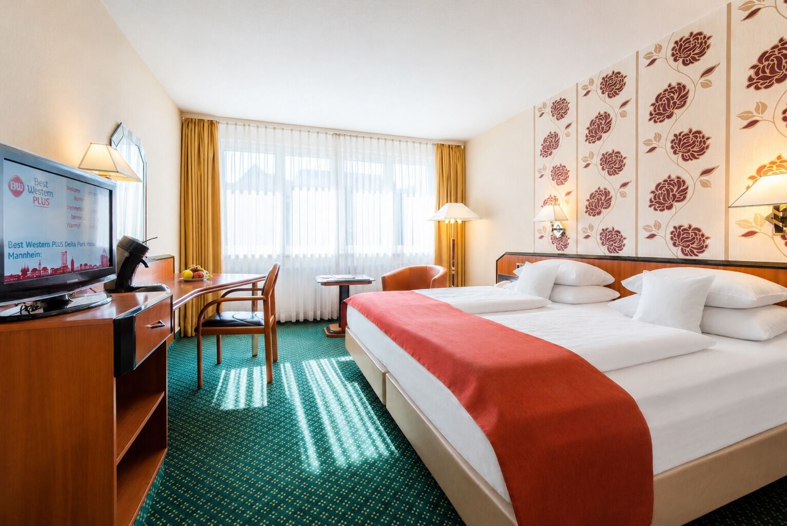 Habitación de hotel con cama, escritorio con computadora, silla, una mesita y televisor mostrando información del hotel.