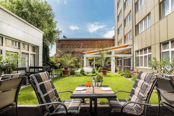 An outdoor seating area with a table and chairs, surrounded by greenery and a building with a brick facade.