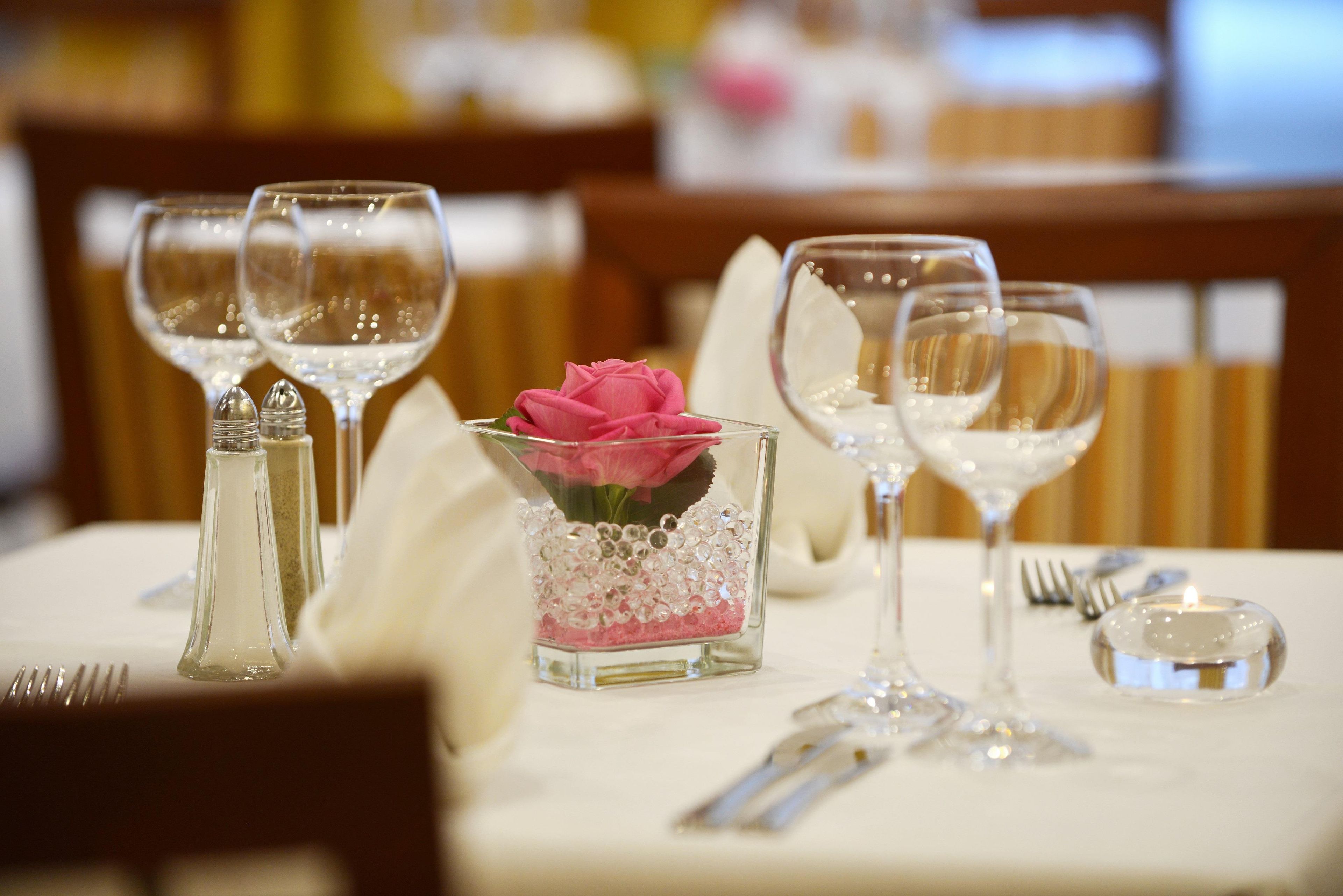 Una mesa dispuesta para comer, con copas de vino, un florero con una flor rosa y cubiertos.