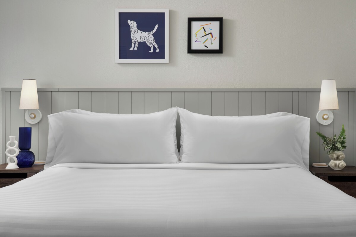 Un lit bien fait, avec du linge de lit blanc, deux lampes fixées au mur et deux œuvres d’art encadrées accrochées au mur.